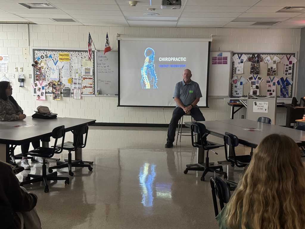 Guest speaker sits before students during a chiropractic workshop, with a skeleton image projected behind him.