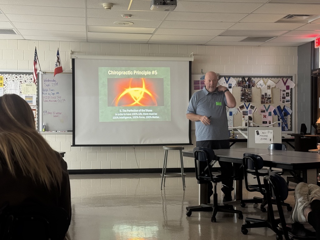 Guest speaker presents a chiropractic principle to students while standing beside a projected classroom slide.