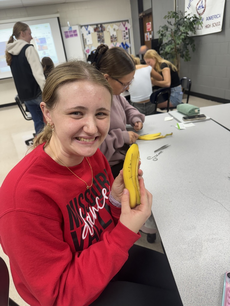 Smiling student holds up a banana while classmates continue a hands-on activity at a classroom table.