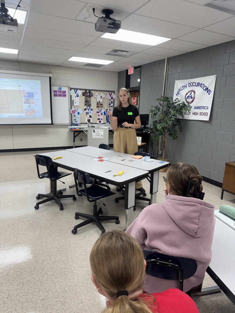 Instructor addresses seated students during a hands-on classroom lesson, with activity materials on the tables.
