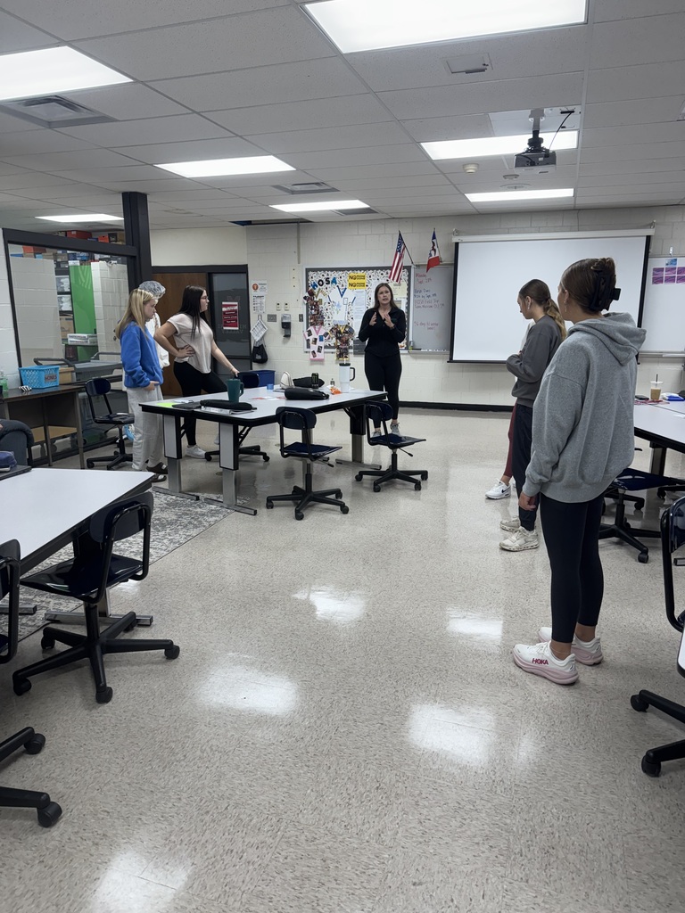 Students stand around a classroom as an instructor demonstrates an activity at the front of the room.