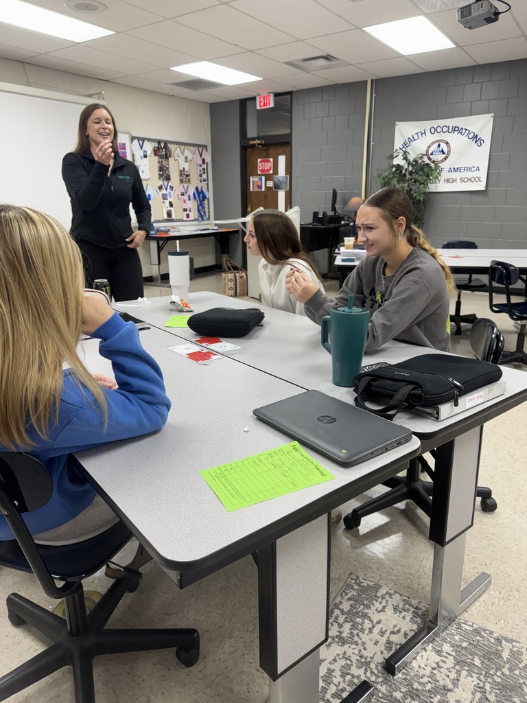 An instructor leads an activity as students seated around classroom tables watch and participate.