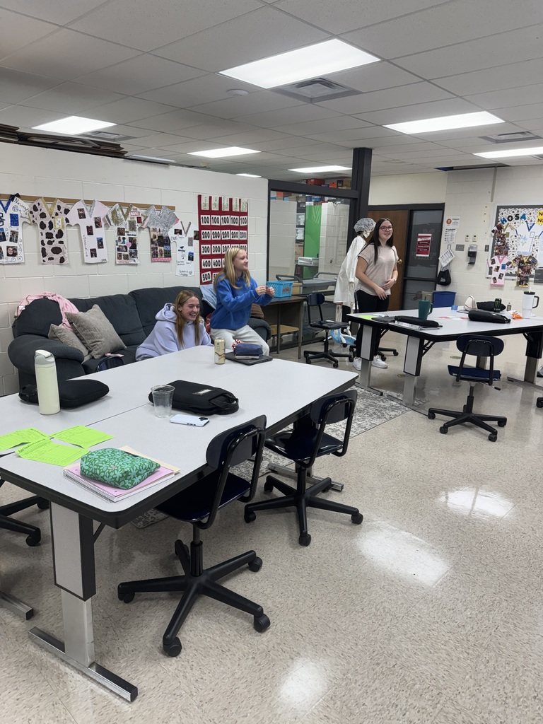 Students smile and interact during a classroom activity while an instructor stands nearby.