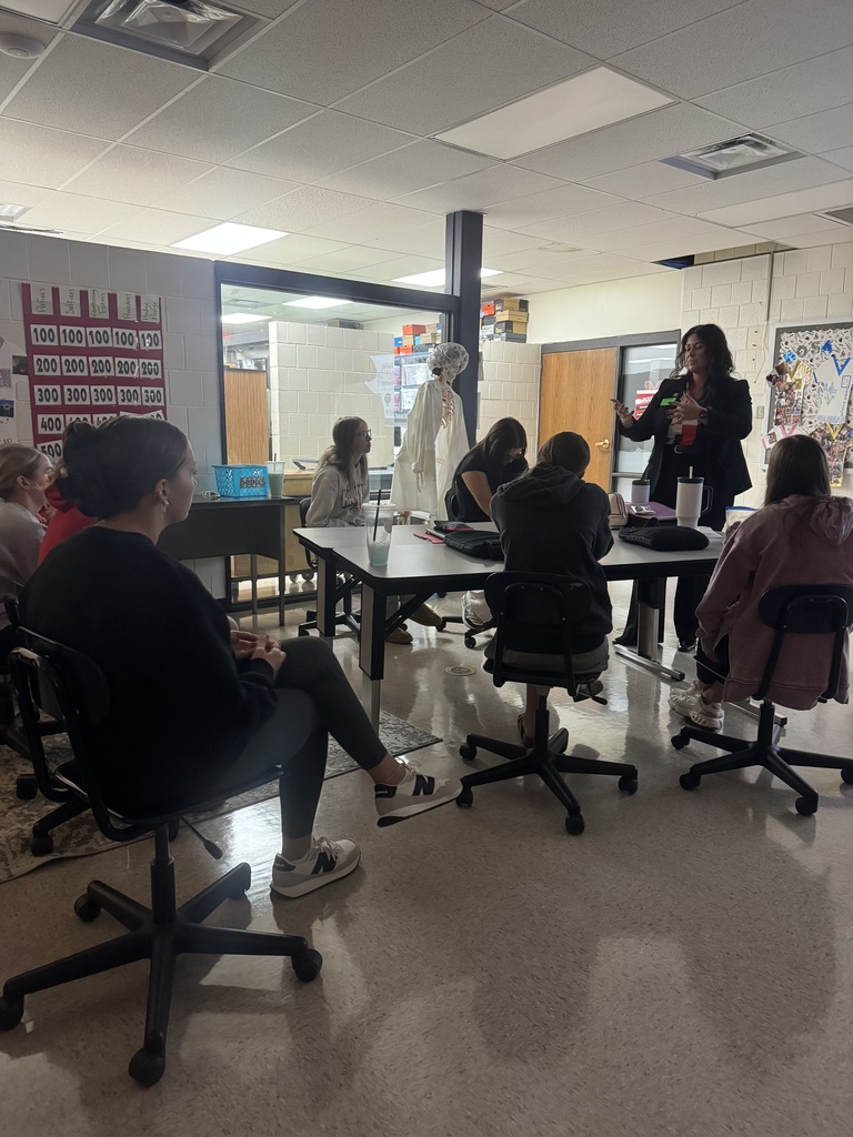 A funeral service professional speaks to students seated around classroom tables, with an anatomical skeleton model nearby.