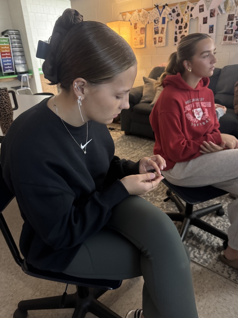 Two students sit in a classroom, one examining a small object while the other listens during a funeral service career presentation.