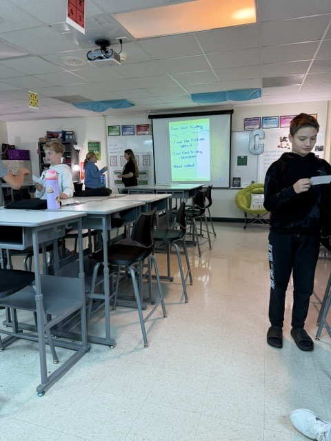 Students participate in a Fun Fact Friday classroom activity while one student reads a paper in the foreground.