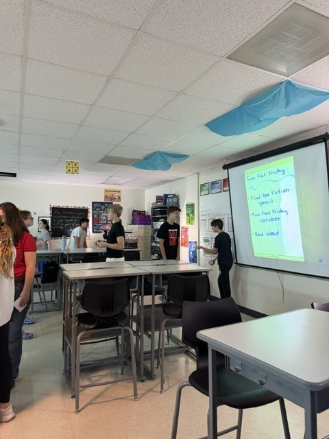 Students move around a classroom during a group activity, with Fun Fact Friday instructions projected on the screen.