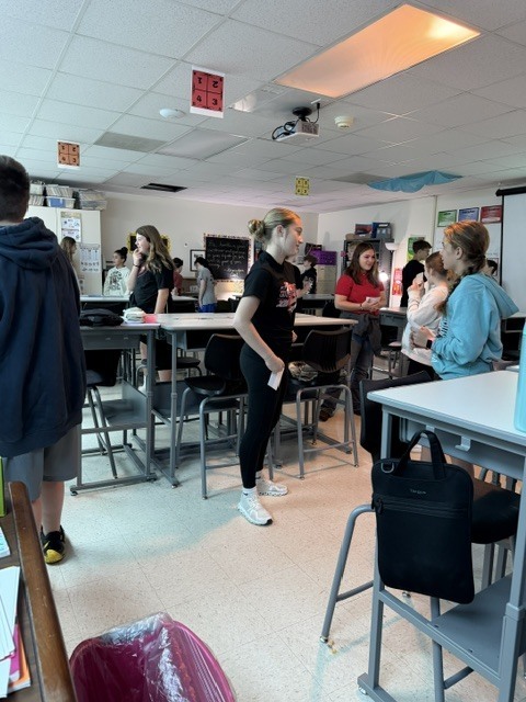 Students stand and socialize around tall tables in a classroom, participating in a group activity.