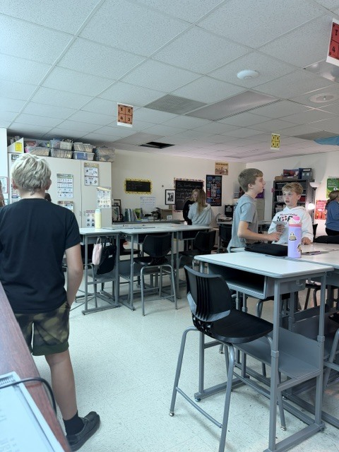 Students converse in small groups around classroom tables during an interactive learning activity.