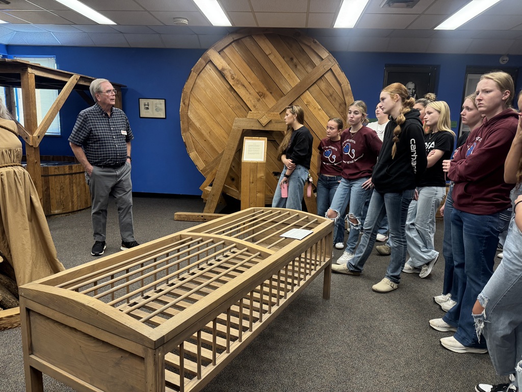 Museum guide speaks to students gathered around historical wooden exhibits, including a large circular structure.