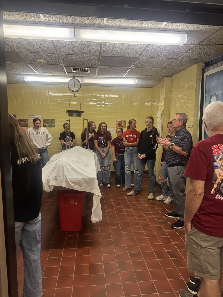 Museum guide explains a historical exhibit to students in a yellow-tiled room featuring a covered table and hanging scale.