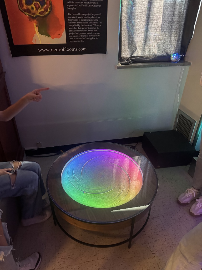 Colorful rainbow light patterns swirl across a circular interactive exhibit as students observe in a dimly lit room.