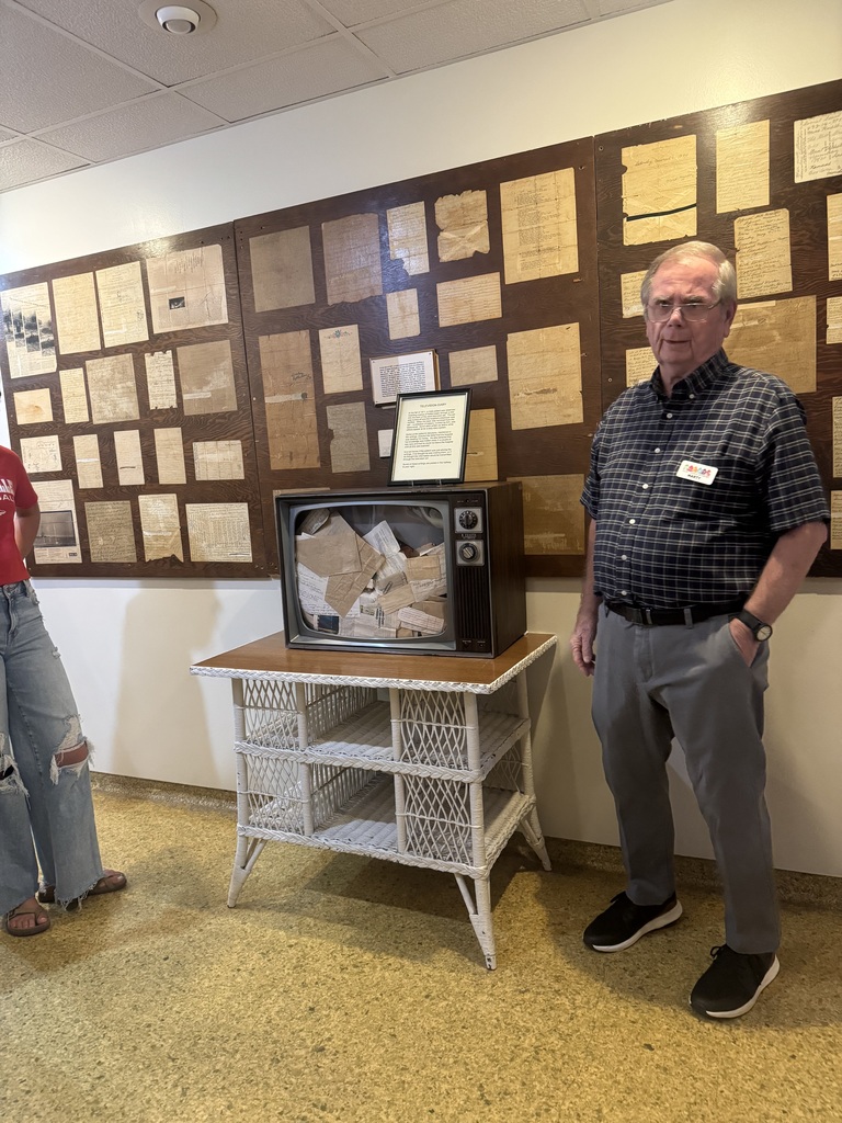 Museum guide stands beside a vintage television filled with historical documents, with archival papers displayed on the wall.