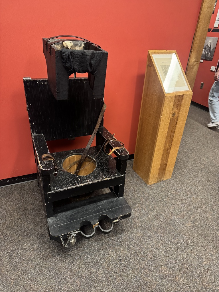 Antique wooden restraint chair with straps and a foot lock displayed beside an informational plaque in a museum.