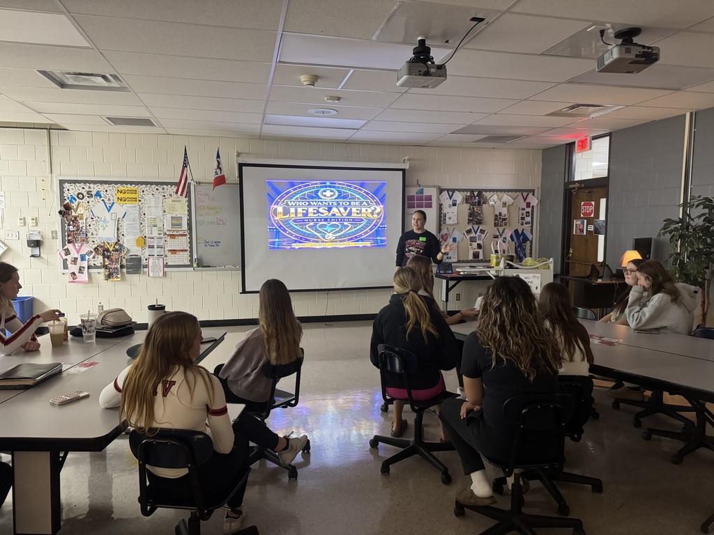 An instructor leads a “Who Wants to Be a Lifesaver?” nursing presentation as students watch from classroom tables.