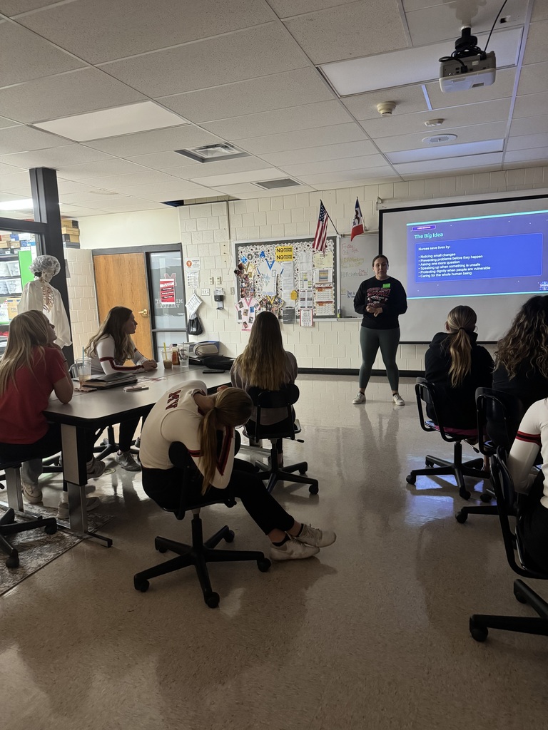 Students listen to a presenter discussing ways nurses save lives, with key points displayed on a classroom projection screen.