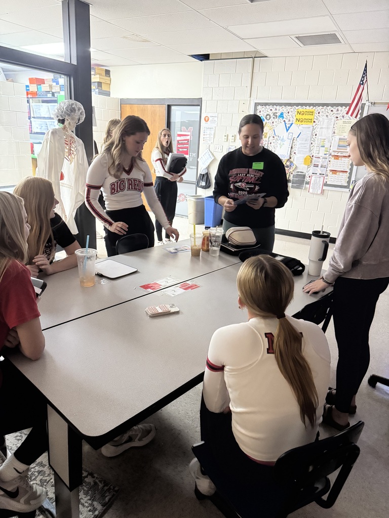 Students in Big Red apparel gather around a classroom table with an instructor, reviewing materials during a group activity.