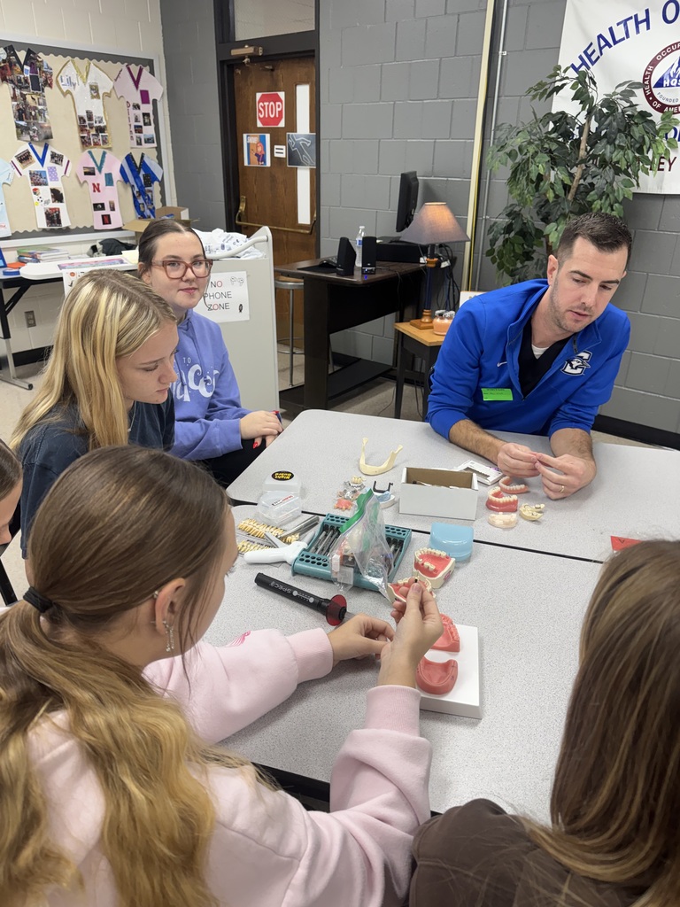 A dental professional demonstrates dental models and instruments to students gathered around a classroom table.