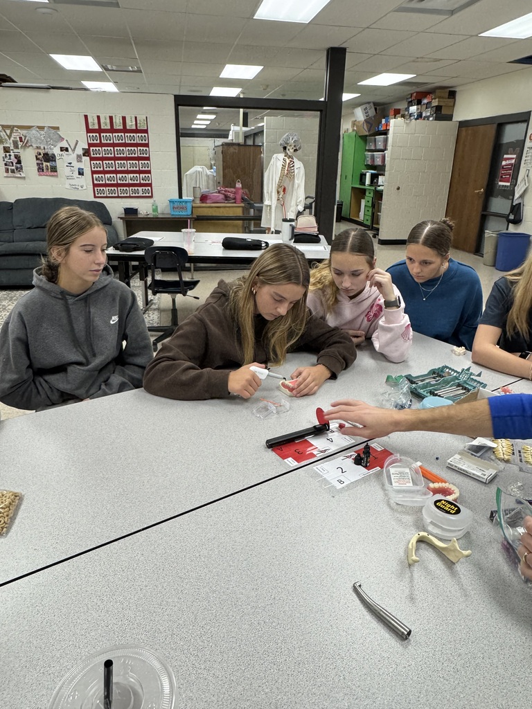 A student practices using a dental instrument on a model of teeth while classmates observe in a classroom.