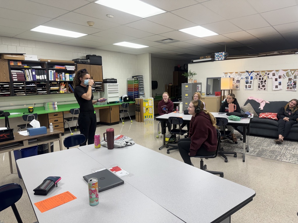 A guest speaker holds a phone while leading a classroom activity as students smile and participate, with one wearing headphones.