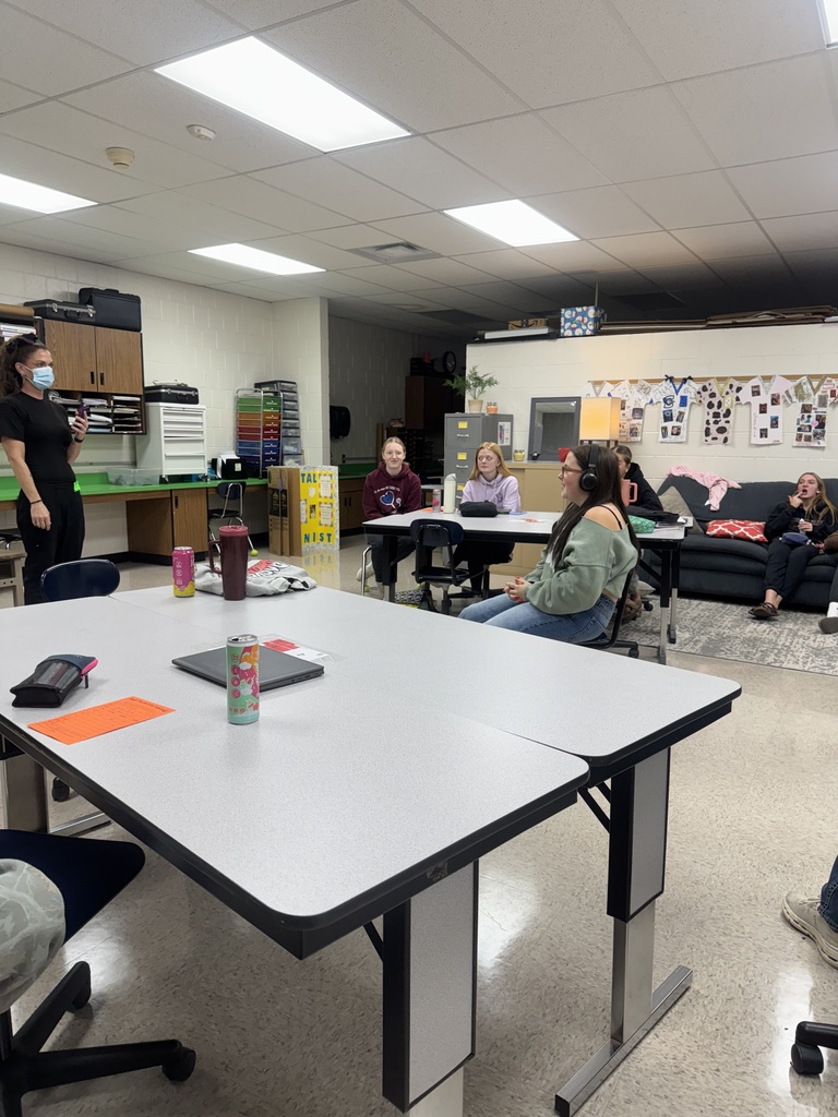 A guest speaker wearing a face mask addresses students during an interactive classroom activity involving headphones.