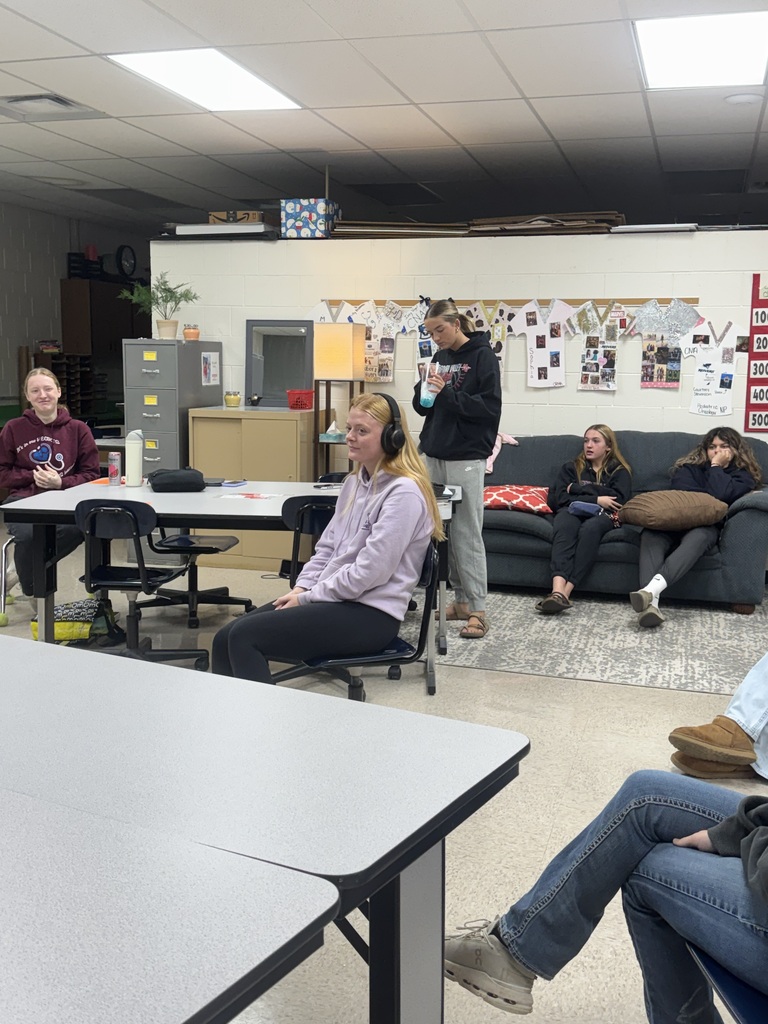 A student in a lavender hoodie wears headphones during a classroom activity as classmates observe from nearby tables and a couch.