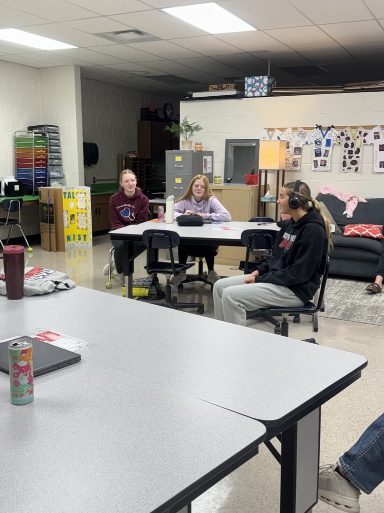 Three students participate in a classroom listening activity, with one student wearing headphones while seated apart from the others.