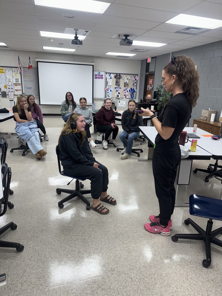 A guest speaker holds a phone toward a seated student wearing headphones as classmates watch the demonstration.