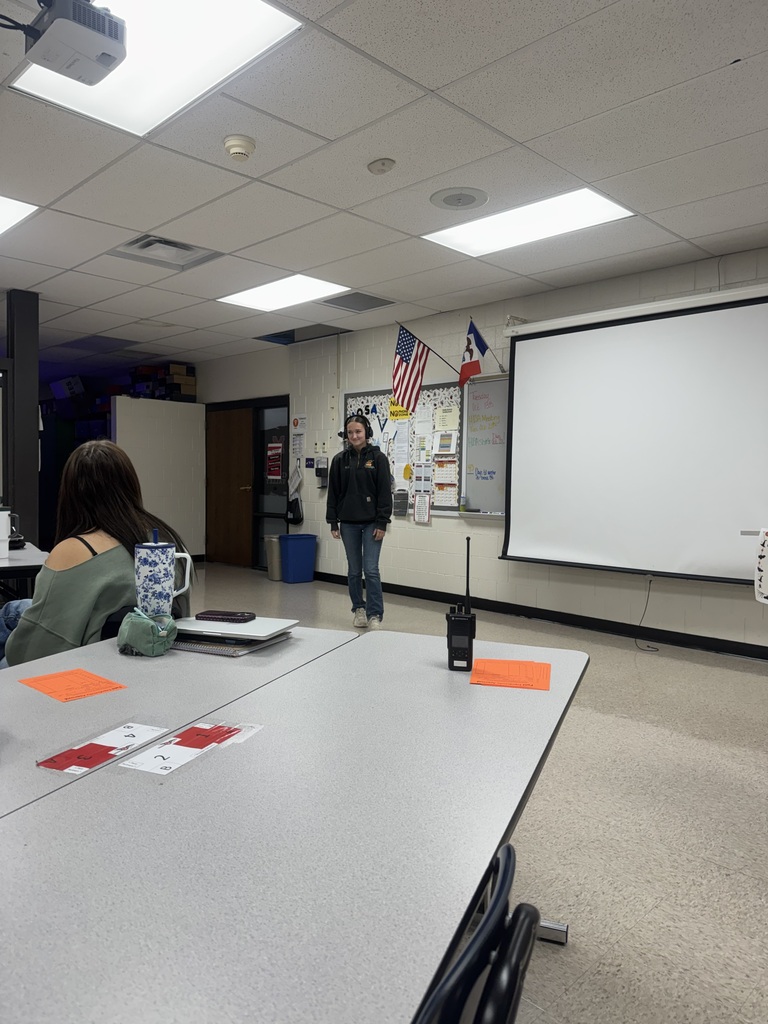 A student wearing headphones stands at the front of a classroom near a projection screen during a group activity.