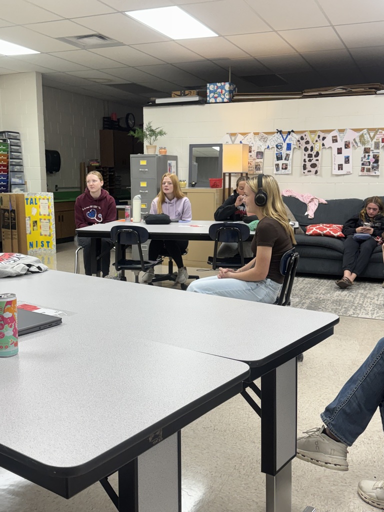 A student wearing headphones sits at the front of a classroom while other students watch and listen from tables and a couch.