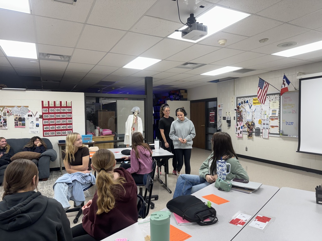 A student wearing headphones stands beside a guest speaker while classmates watch from their seats during an interactive activity.