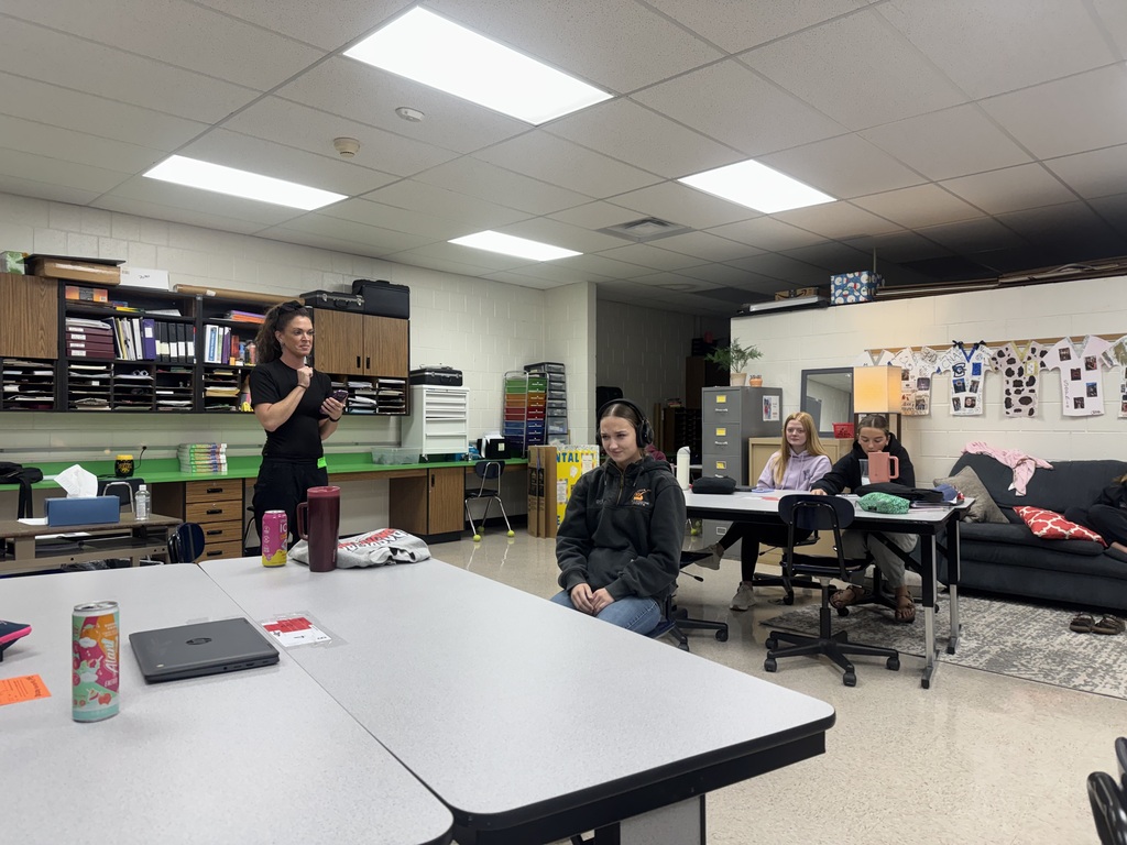 A guest speaker leads an interactive listening activity while a student wearing headphones sits at the front of the classroom.