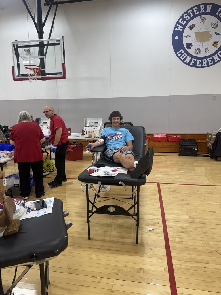 A student in a blue shirt smiles while donating blood, with Red Cross staff and equipment nearby in the school gym.