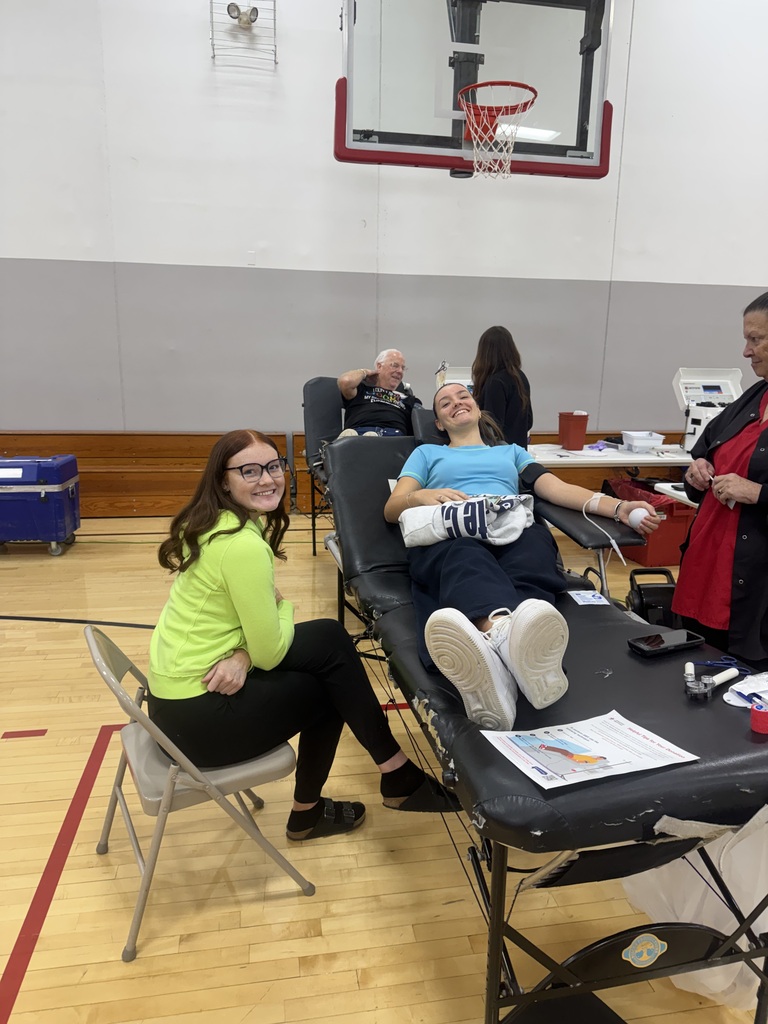 A smiling student donates blood while a woman in a bright green shirt sits beside her and a staff member stands nearby.