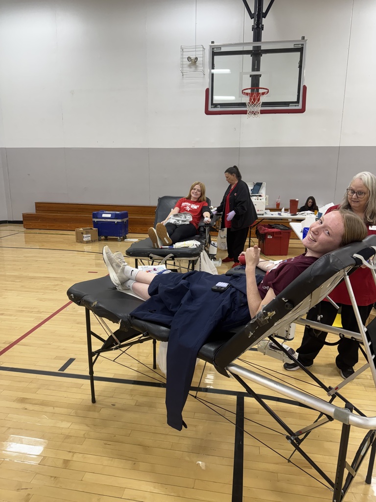 A smiling student reclines on a blood donation cot while another donor and Red Cross staff work nearby in a school gym.
