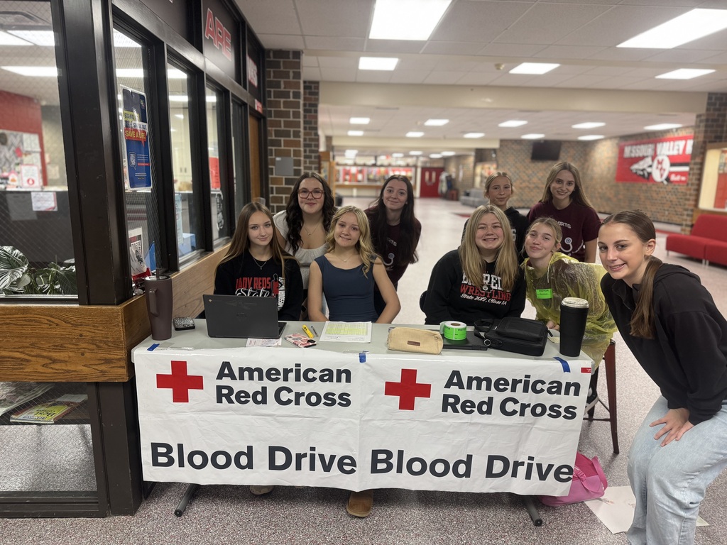 Nine students pose behind an American Red Cross Blood Drive registration table in a school hallway.