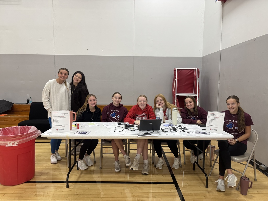 Eight student volunteers smile behind a registration table with laptops and paperwork at a school blood drive.