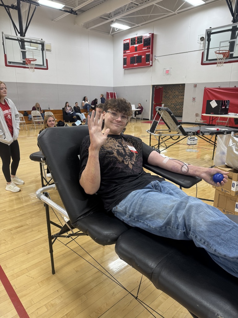 A smiling student waves while donating blood on a reclining cot in a school gymnasium, holding a blue stress ball.