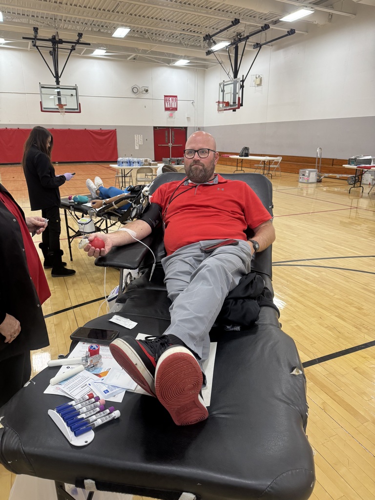 A man in a red polo shirt donates blood while reclining on a cot in a school gym, holding a red stress ball.