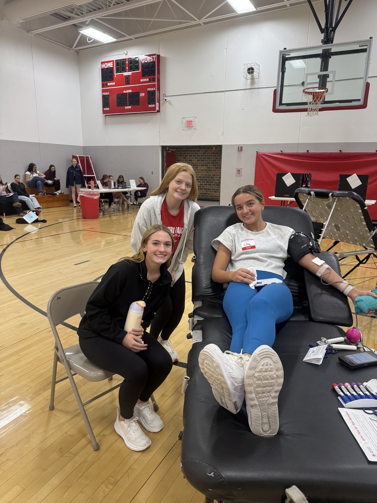 Three smiling students gather around a classmate donating blood on a cot during a school blood drive.