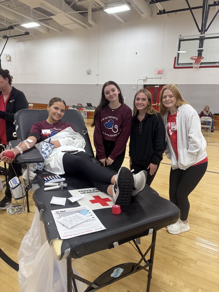 A smiling student donates blood while reclining on a cot, surrounded by three supportive classmates at a school blood drive.