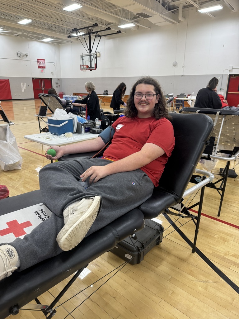 A student in a red shirt smiles while donating blood on a cot, holding a green stress ball in the school gymnasium.