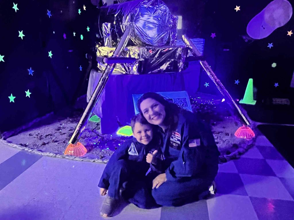 A teacher and student wearing NASA costumes and sitting in front of a mock-up of a moon landing craft