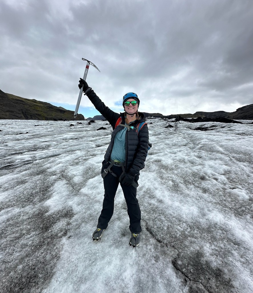 A teacher in a parka holding up a pickaxe while standing on a glacier