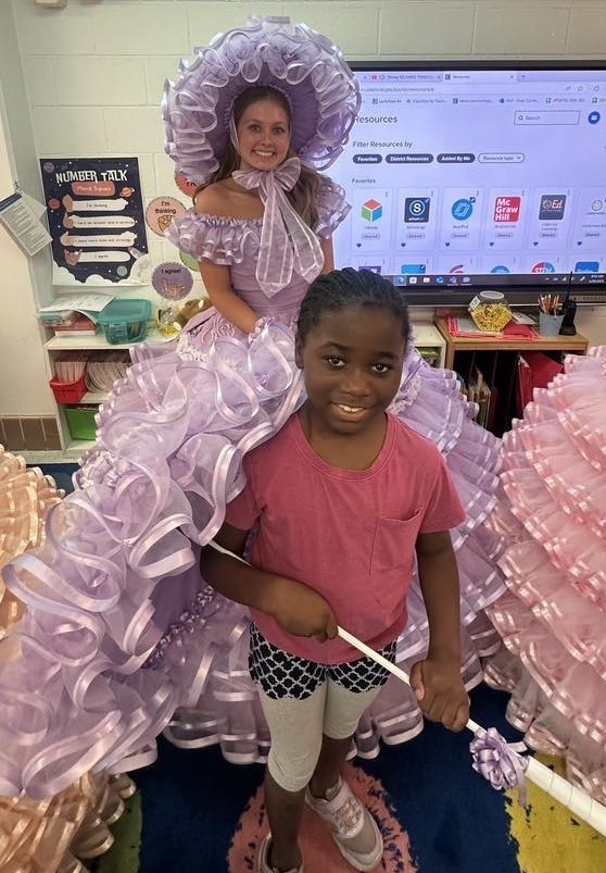An Azalea Trail Maid in a lavendar hoop dress stands behind and elementary student who is holding her parasol