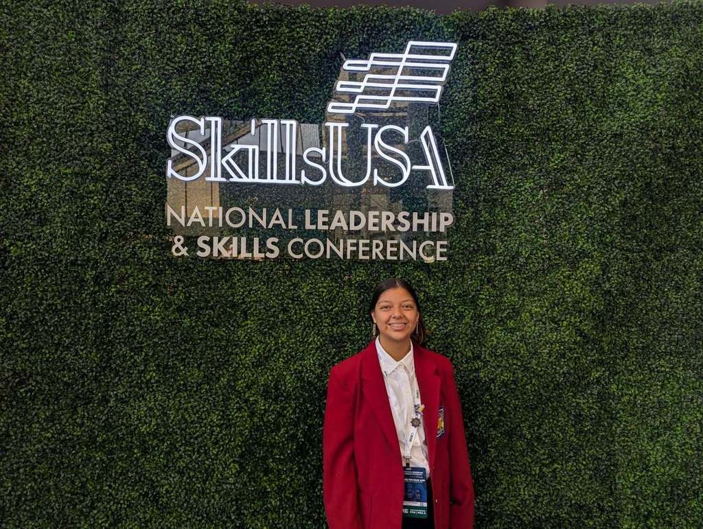 A photo of Mia Lopez, wearing a red blazer and standing in front of a sign that says: "Skills USA National Leadership & Skills Conference"