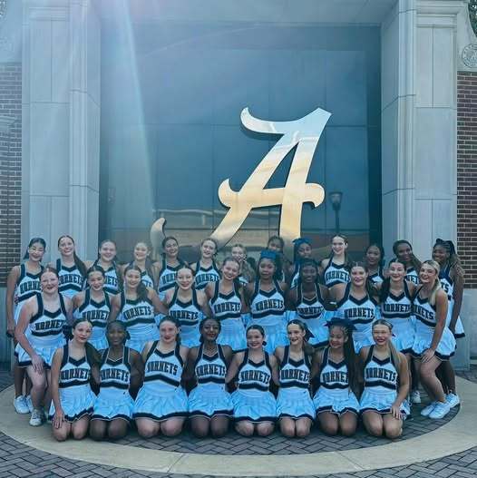 A group photo of the Baker cheerleaders in front of a large window with the letter "A" logo of the University of Alabama on it