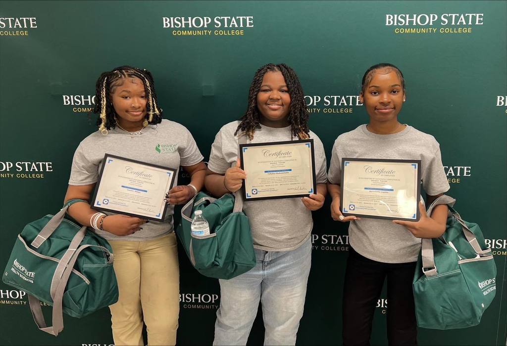 Three girls holding certificates and carrying green duffel bags, standing before a backdrop with the Bishop State logo on it