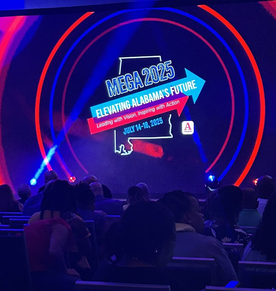 A darkened ballroom, bathed in blue light, with the logo of the Mega Conference on a screen: "Mega 2025: Elevating Alabama's Future"