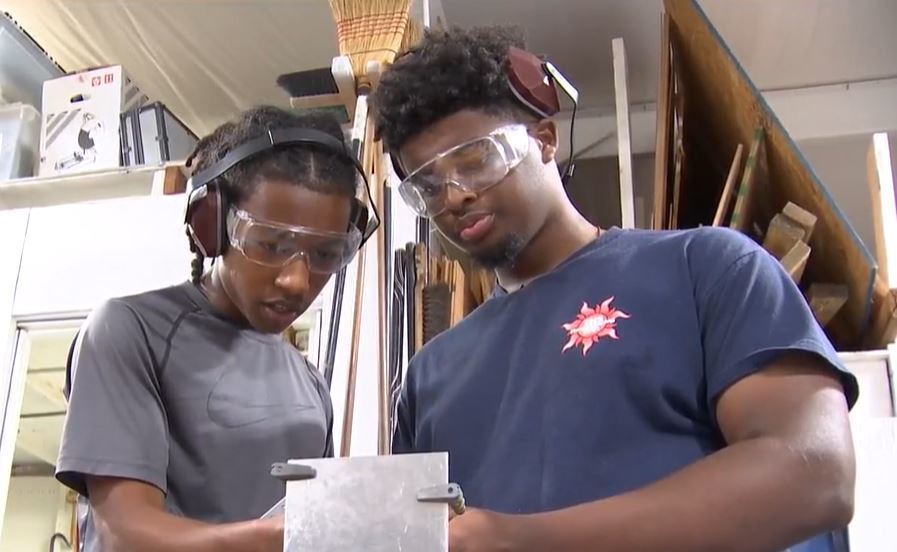 Two students in safety goggles and ear protectors working with a piece of metal
